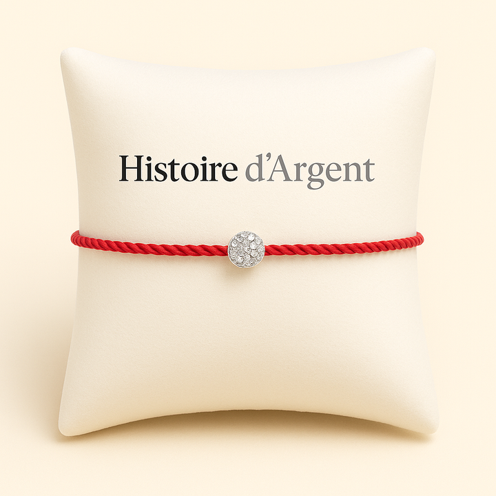 Bracelet en fil rouge tressé avec perle zircon centrale, présenté sur coussin blanc élégant, logo Histoire d’Argent en signature de marque.